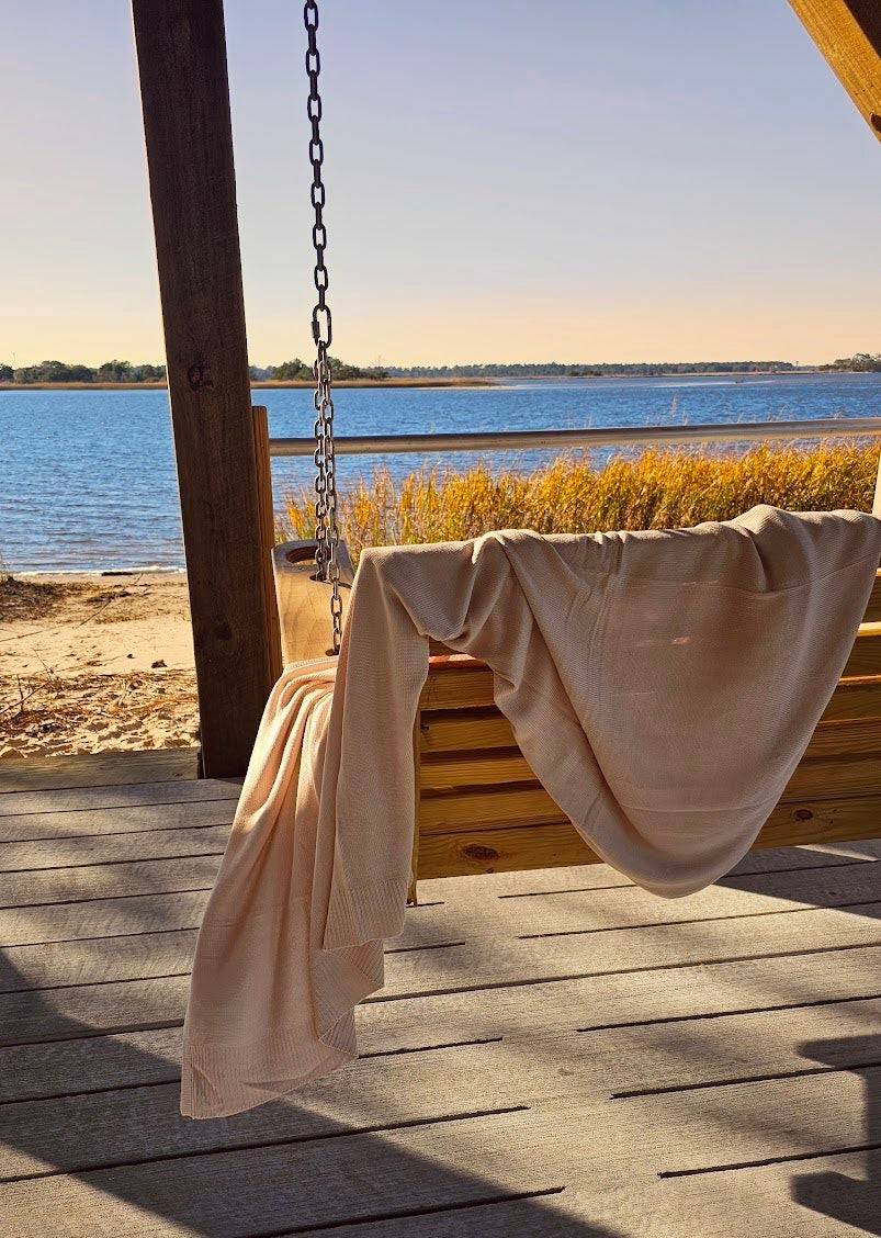 The drift throw in beige almond color over porch swing overlooking ocean at sunset, lightweight throw over rocking chair blowing in the beach breeze, oversized throw, travel throw, outdoor throw blanket, easy clean throw, durable bedding