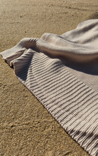 The drift throw in beige almond color laying in the sand, sunlight sunset view lightweight throw over rocking chair blowing in the beach breeze, oversized throw, travel throw, outdoor throw blanket, easy clean throw, durable bedding