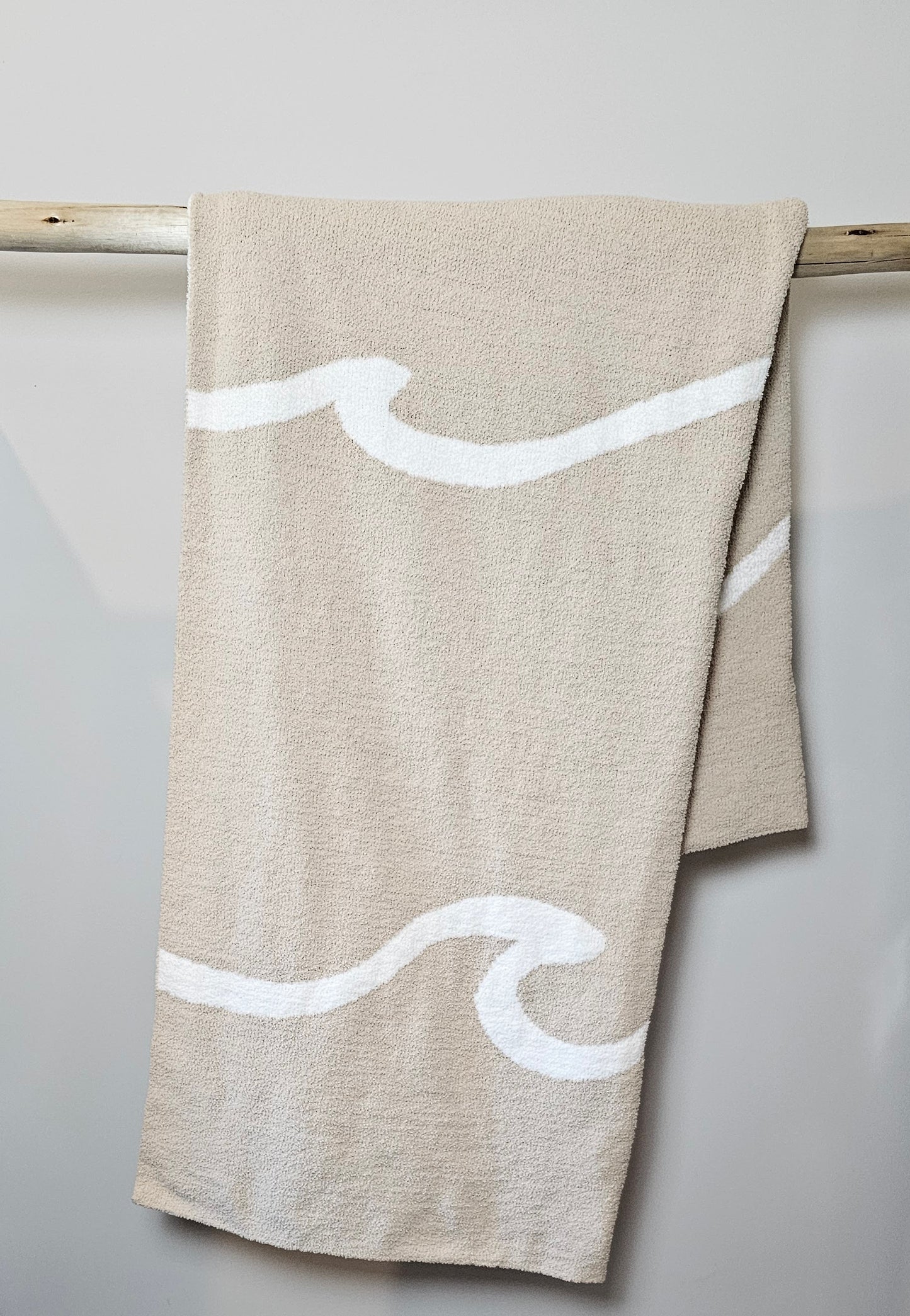 Wave nest throw blanket artfully draped over driftwood, featuring its ocean-inspired design. Soft and stylish Wave throw hanging on driftwood, embodying beachside comfort and elegance. showcasing its Double sided design with sand beige and white

