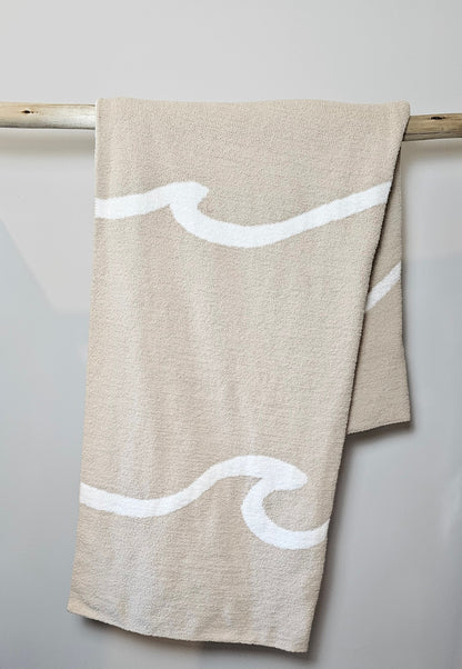 Wave nest throw blanket artfully draped over driftwood, featuring its ocean-inspired design. Soft and stylish Wave throw hanging on driftwood, embodying beachside comfort and elegance. showcasing its Double sided design with sand beige and white

