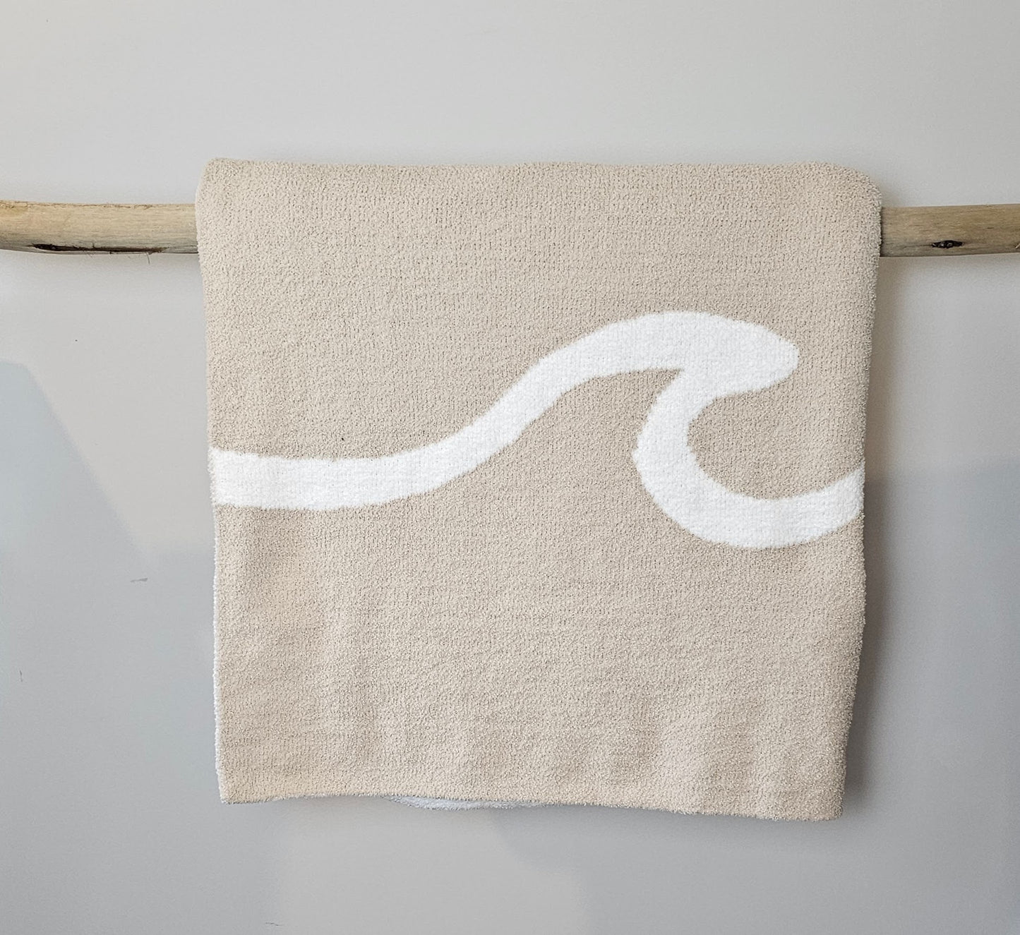 Wave nest throw blanket artfully draped over driftwood, featuring its ocean-inspired design. Soft and stylish Wave throw hanging on driftwood, embodying beachside comfort and elegance. showcasing its Double sided design with sand beige and white

