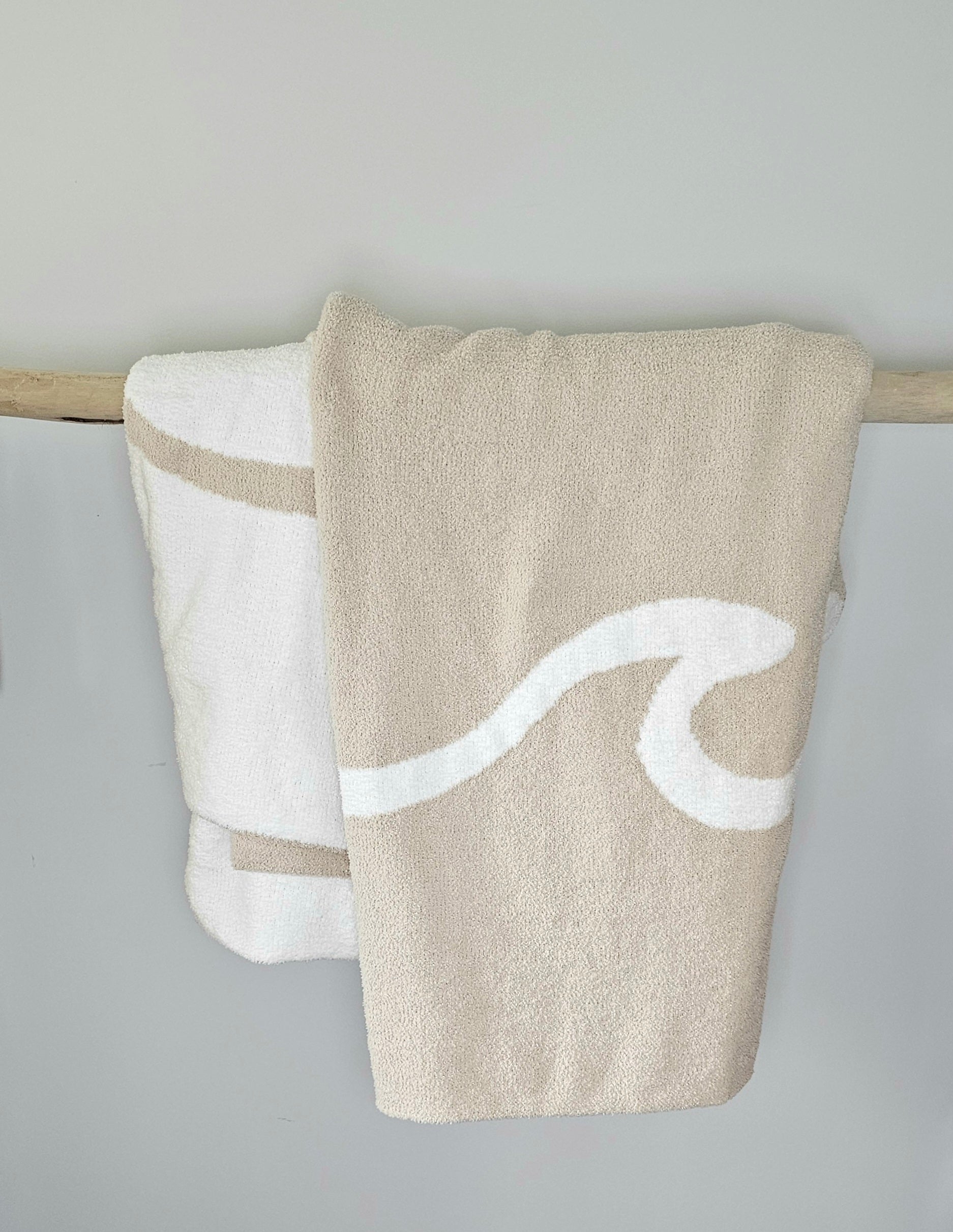 Wave nest throw blanket artfully draped over driftwood, featuring its ocean-inspired design. Soft and stylish Wave throw hanging on driftwood, embodying beachside comfort and elegance. showcasing its Double sided design with sand beige and white

