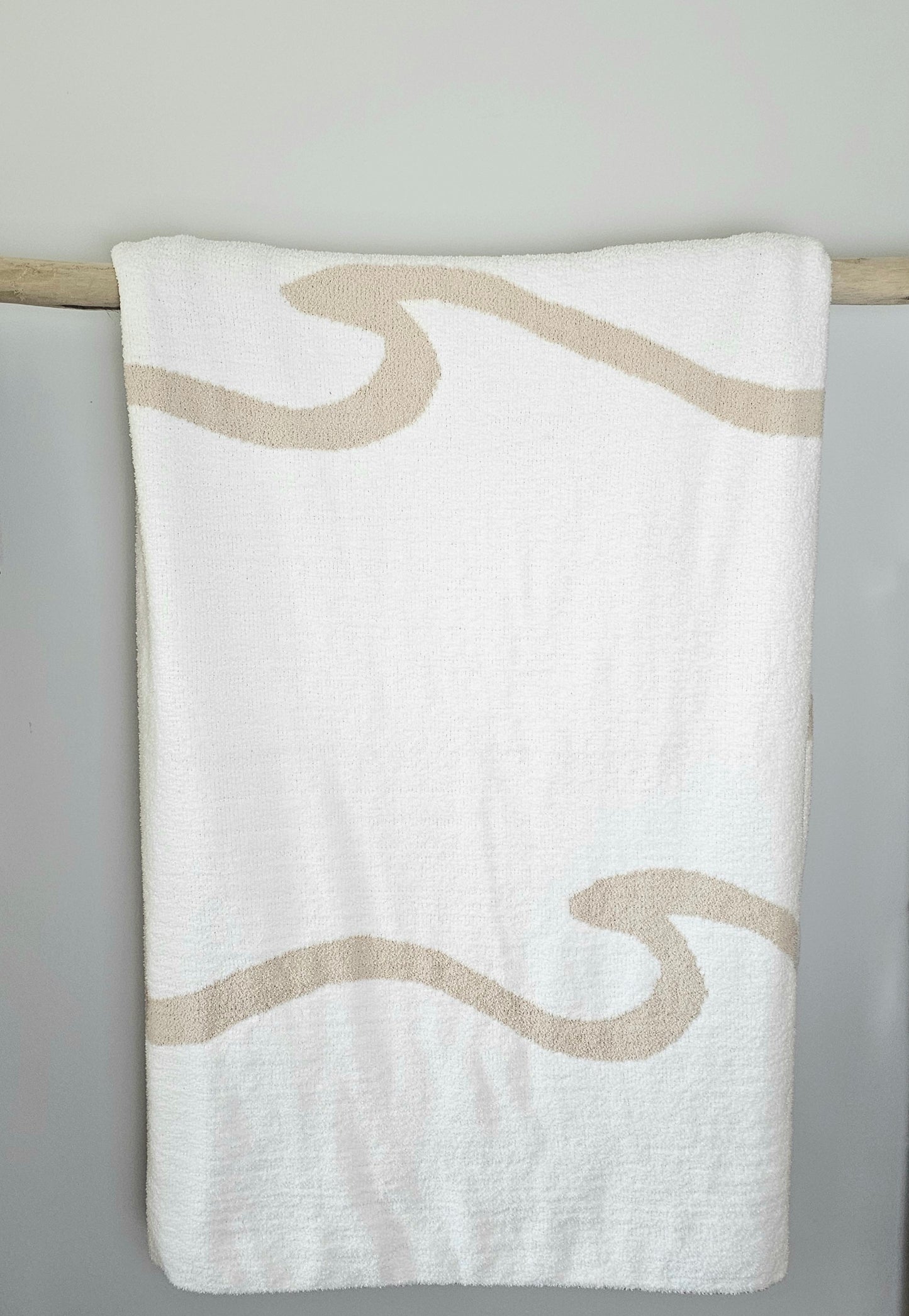 Wave nest throw blanket artfully draped over driftwood, featuring its ocean-inspired design. Soft and stylish Wave throw hanging on driftwood, embodying beachside comfort and elegance. showcasing its Double sided design with sand beige and white

