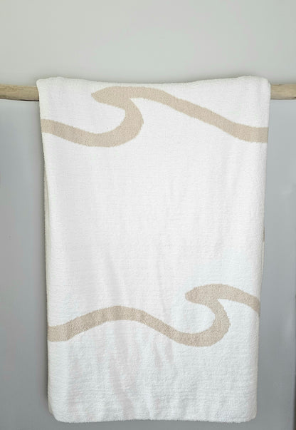 Wave nest throw blanket artfully draped over driftwood, featuring its ocean-inspired design. Soft and stylish Wave throw hanging on driftwood, embodying beachside comfort and elegance. showcasing its Double sided design with sand beige and white

