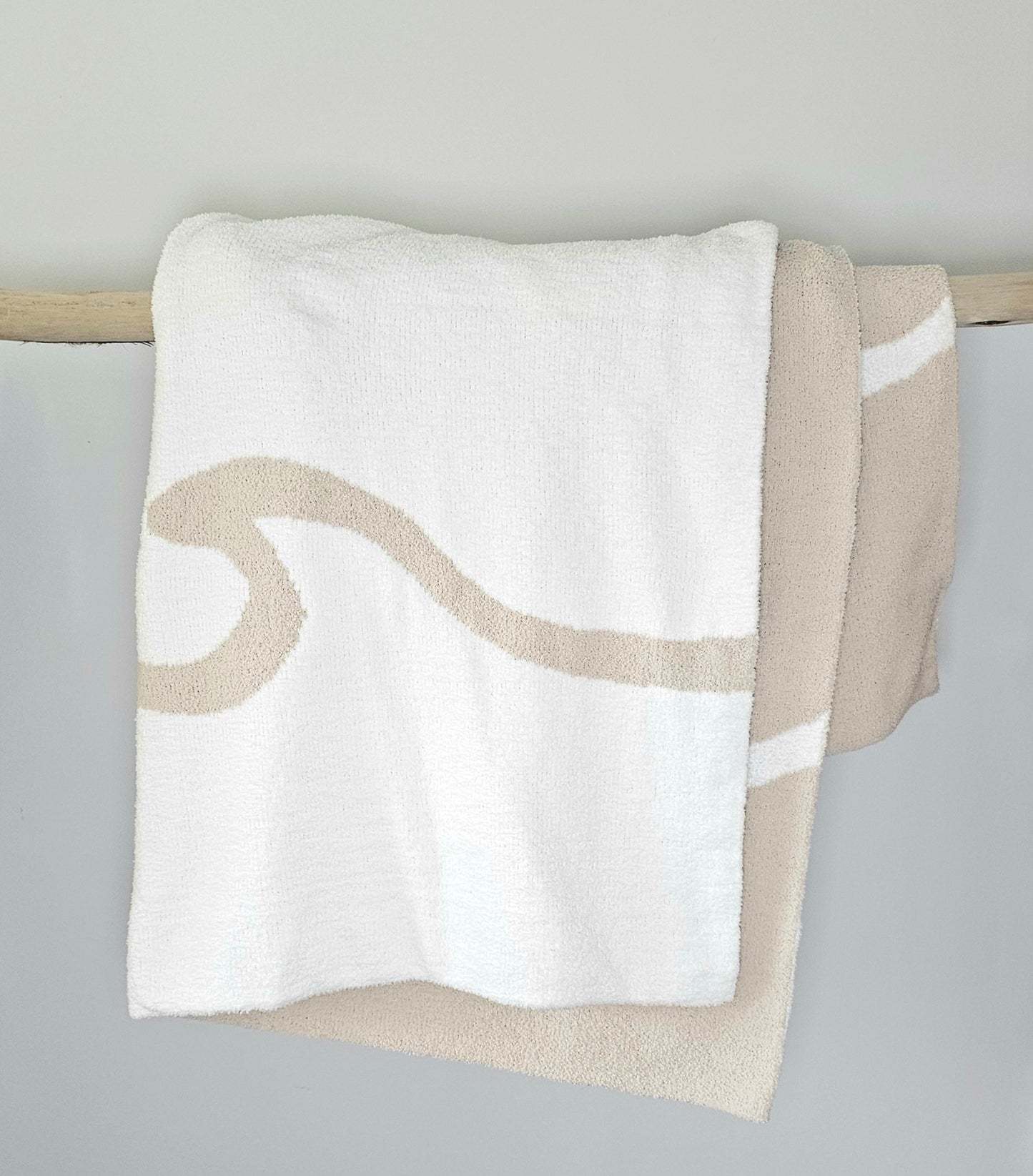 Wave nest throw blanket artfully draped over driftwood, featuring its ocean-inspired design. Soft and stylish Wave throw hanging on driftwood, embodying beachside comfort and elegance. showcasing its Double sided design with sand beige and white

